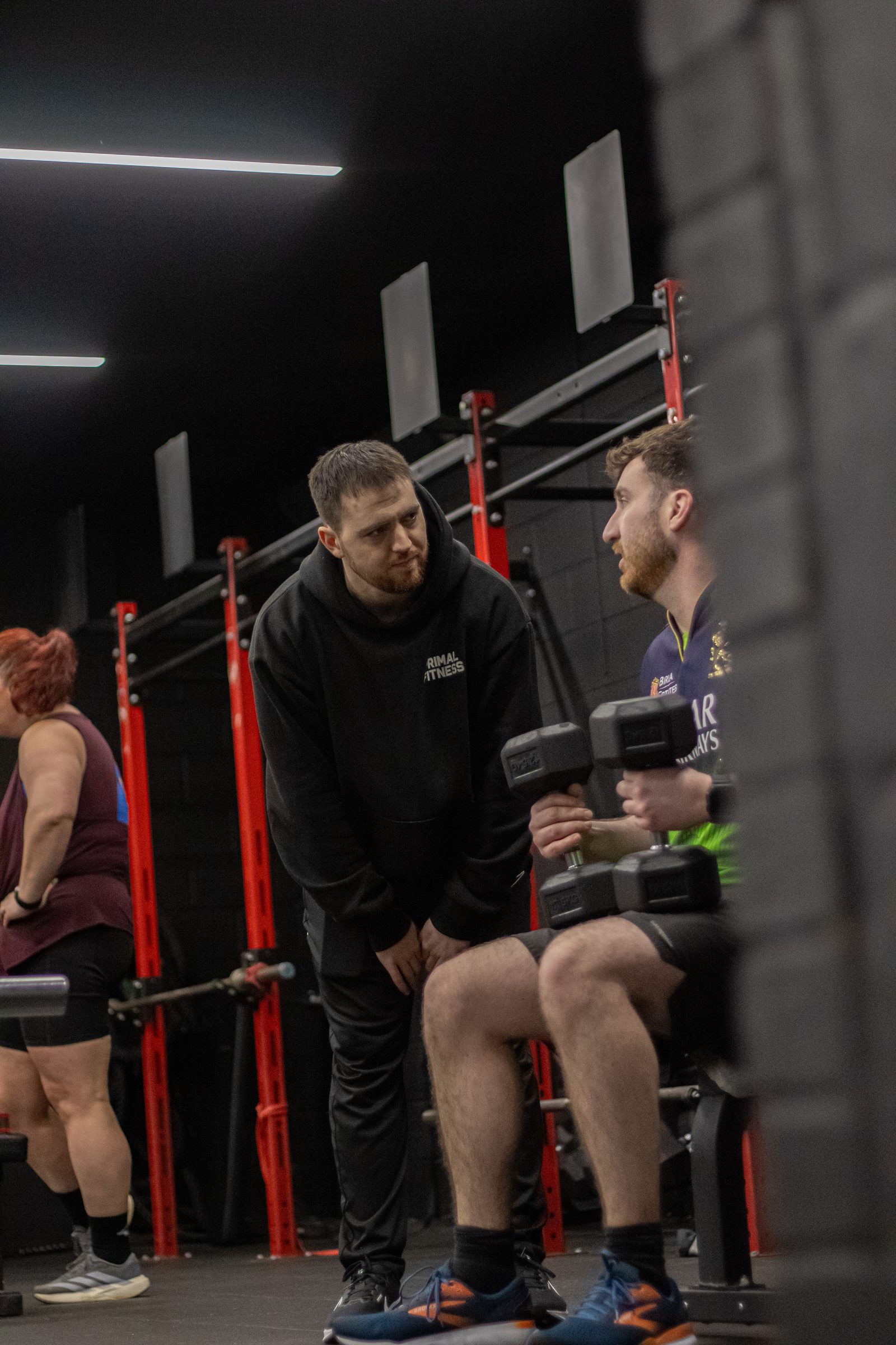 A Primal coach crouched beside a member pressing dumbbells, by the red rigs